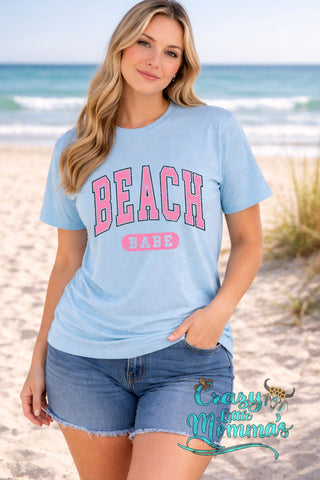 A woman on a beach wearing a light blue "Beach Babe" tee with pink lettering and denim shorts.