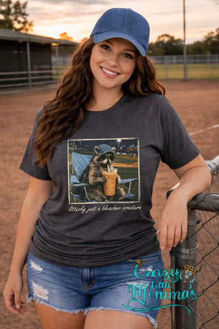 A woman in a dark gray tee with a graphic of a raccoon in a baseball cap & sunglasses, drinking coffee.