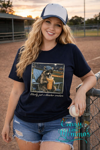 Woman in a navy tee with a raccoon in a baseball cap and sunglasses drinking iced coffee.