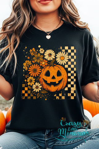 A woman wears a black Checkered Pumpkin Tee with a smiling jack-o'-lantern and fall flowers.
