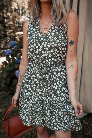 Woman wearing an olive floral mini dress with a tie waist and white daisy print, holding a brown purse.