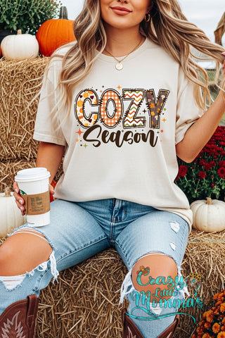 Woman in a cream "Cozy Season" graphic tee with plaid and autumn patterns, holding coffee on hay bales.