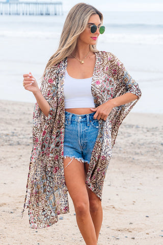 Woman on beach wearing a Desert Bloom Kimono with a bohemian floral pattern over a white crop top and denim shorts.