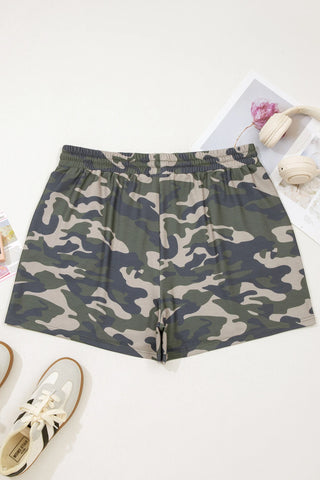 Desert Trail Camo Shorts with an elastic waistband, laid flat on a white surface with sneakers and headphones.
