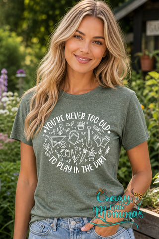 Woman in a green tee with "You're Never Too Old To Play In The Dirt" and gardening tools graphic.