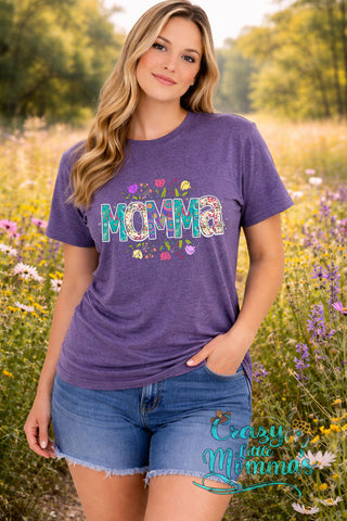 Woman in a field wearing a purple "Momma" tee with floral and paisley patterns, and denim shorts.