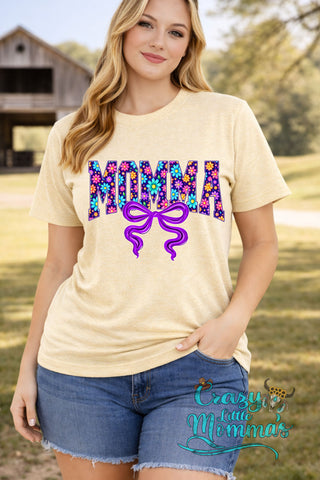 A woman models a beige tee with "MOMMA" in colorful floral print and a purple bow graphic.