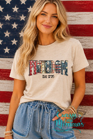 Blonde woman in a beige "Freedom" tee with an American flag design, standing against a wooden flag backdrop.