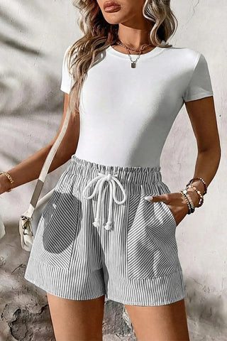 A woman wearing Fresh Linen Stripe Shorts with a white tee, holding a white bag, hand in pocket.