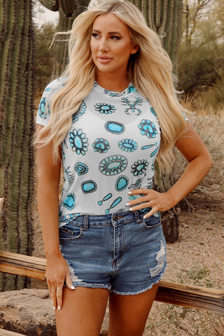 Blonde woman in a white Turquoise Concho Tee with blue concho designs, paired with denim shorts.