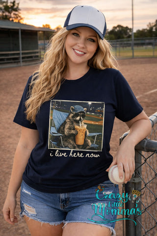 Blonde woman in a navy tee with a raccoon in sunglasses and a baseball cap drinking iced coffee.
