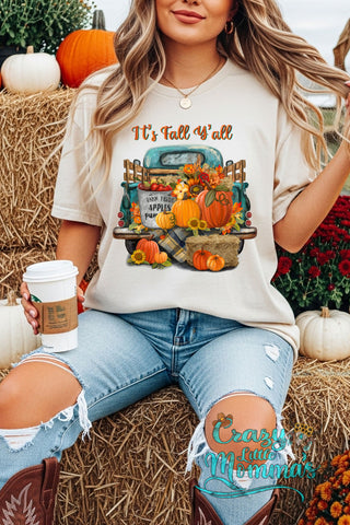 Woman in a cream "It's Fall Y'all" tee with a vintage truck full of pumpkins and fall decor, holding coffee.