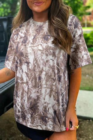 A woman wears an earthy camo crewneck tee with a brown and white leaf pattern.