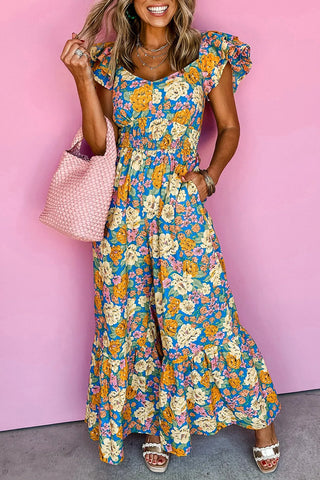 Woman smiling, wearing a blue floral smocked jumpsuit with yellow and pink flowers, holding a pink woven bag.