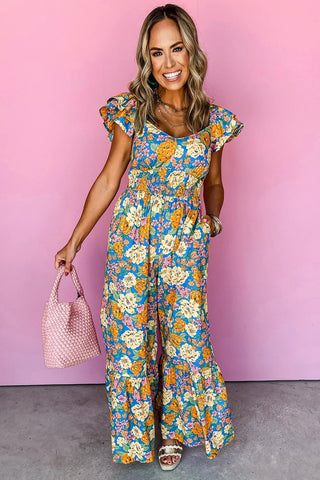 Smiling woman in a blue floral smocked jumpsuit with ruffle sleeves, holding a pink woven bag.