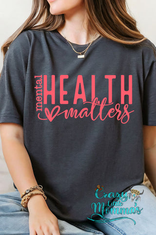 A woman wears a dark gray Mental Health Awareness Tee with "mental HEALTH matters" in pink text.