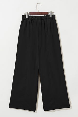 Black wide-leg pants with an elastic waistband and vertical seams, hanging against a white wall.
