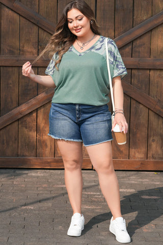 Woman wearing a Prairie Camo Dreams Top, a sage green tee with camo print sleeves, denim shorts, and white sneakers.