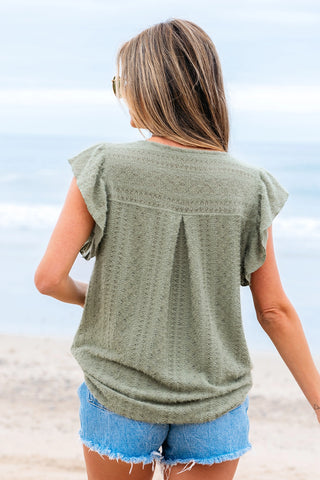 Woman from behind wearing a sage ruffle button top with textured fabric and short sleeves, standing on a beach.