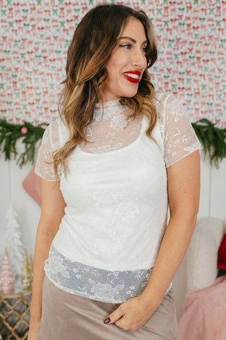 Woman in a white floral lace mockneck top with short sleeves, smiling and looking to the right.