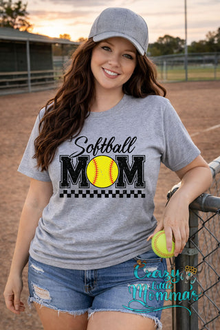 Woman in a grey "Softball Mom" tee with a yellow softball graphic, grey cap, and denim shorts at a ball field.