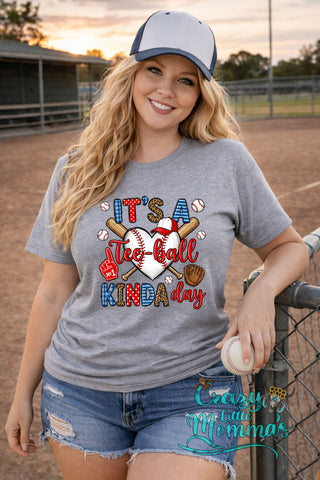 Woman in a gray tee with "It's a Tee-ball Kinda Day" graphic, featuring a baseball heart, bats, glove, and foam finger.