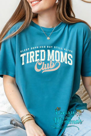 Woman wearing a teal "Tired Moms Club" tee with "Sleep Deprived But Still Alive" text.