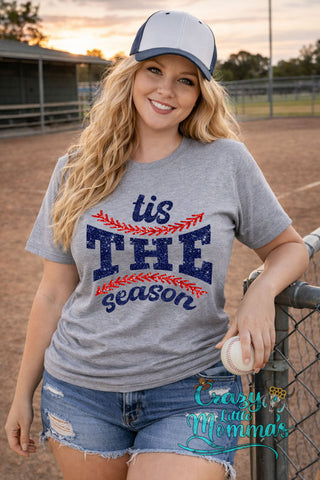 A smiling woman in a grey "Tis The Season" baseball tee with blue glitter text and red baseball stitching.