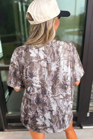 Back view of a woman wearing an earthy camo crewneck tee with a brown and white leaf pattern.
