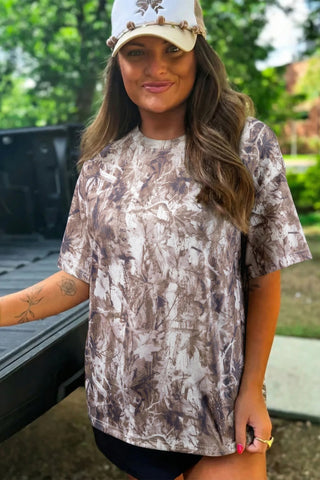 Woman wearing a Wildwood Camo Tee with an earthy camo pattern, a white hat, and black shorts.