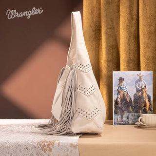 Beige Wrangler hobo bag with studded chevron details and long fringe, sitting on a cowhide rug.