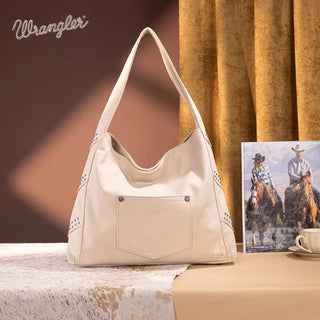 Beige Wrangler studded fringe hobo bag with a front pocket, on a cowhide rug next to a Western-themed picture.