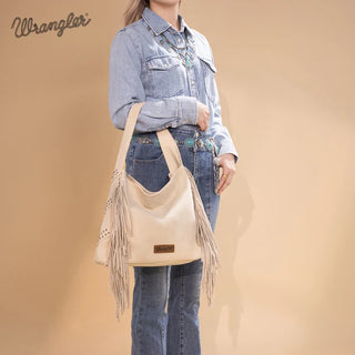 Woman in denim shirt and jeans holds a beige Wrangler studded fringe hobo bag.