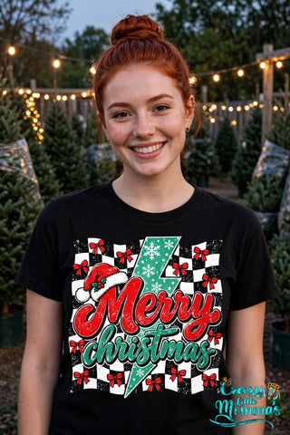 Woman wearing a black tee with the "Merry Christmas" design — red and green script over a black-and-white checkerboard with a green lightning bolt, a Santa hat, and red bows — Crazy Little Momma's logo in the corner.