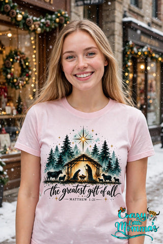 Woman wearing a light pink tee with "The Greatest Gift of All — Matthew 1:21" watercolor nativity design — Crazy Little Momma's logo in the corner.