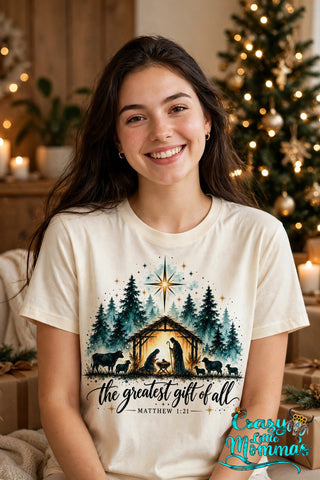 Woman wearing a natural tee with "The Greatest Gift of All — Matthew 1:21" watercolor nativity design — Crazy Little Momma's logo in the corner.