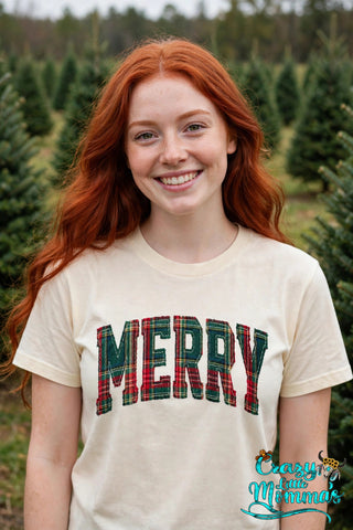 Woman wearing a natural tee with "MERRY" in red and green Christmas plaid letters in a small-town Christmas setting — Crazy Little Momma's logo in the corner.