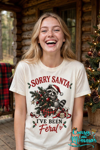 Woman wearing a natural tee with the "Sorry Santa, I've Been Feral" design — a raccoon in a Santa hat tangled in a Christmas tree — Crazy Little Momma's logo in the corner.