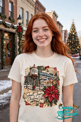 Woman wearing a natural tee with the "Small Town Christmas Vibes" design — a water tower, a snowy small town, and a big red poinsettia — Crazy Little Momma's logo in the corner.