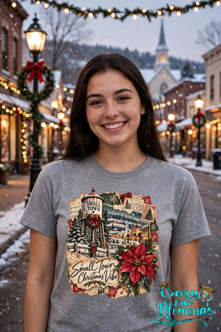 Woman wearing a sport gray tee with the "Small Town Christmas Vibes" design — a water tower, a snowy small town, and a big red poinsettia — Crazy Little Momma's logo in the corner.