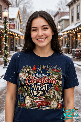 Woman wearing a navy tee with the "Christmas in West Virginia" design, standing by a Christmas tree on a small-town street with her boots in the snow — Crazy Little Momma's logo in the corner.