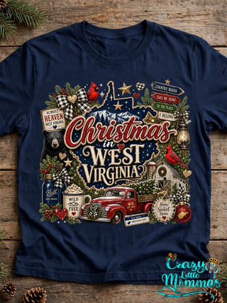 Navy tee with the "Christmas in West Virginia" design laid flat on rustic wood with pine sprigs — the design shows the state outline, a black bear, cardinals, a red truck, and a barn.